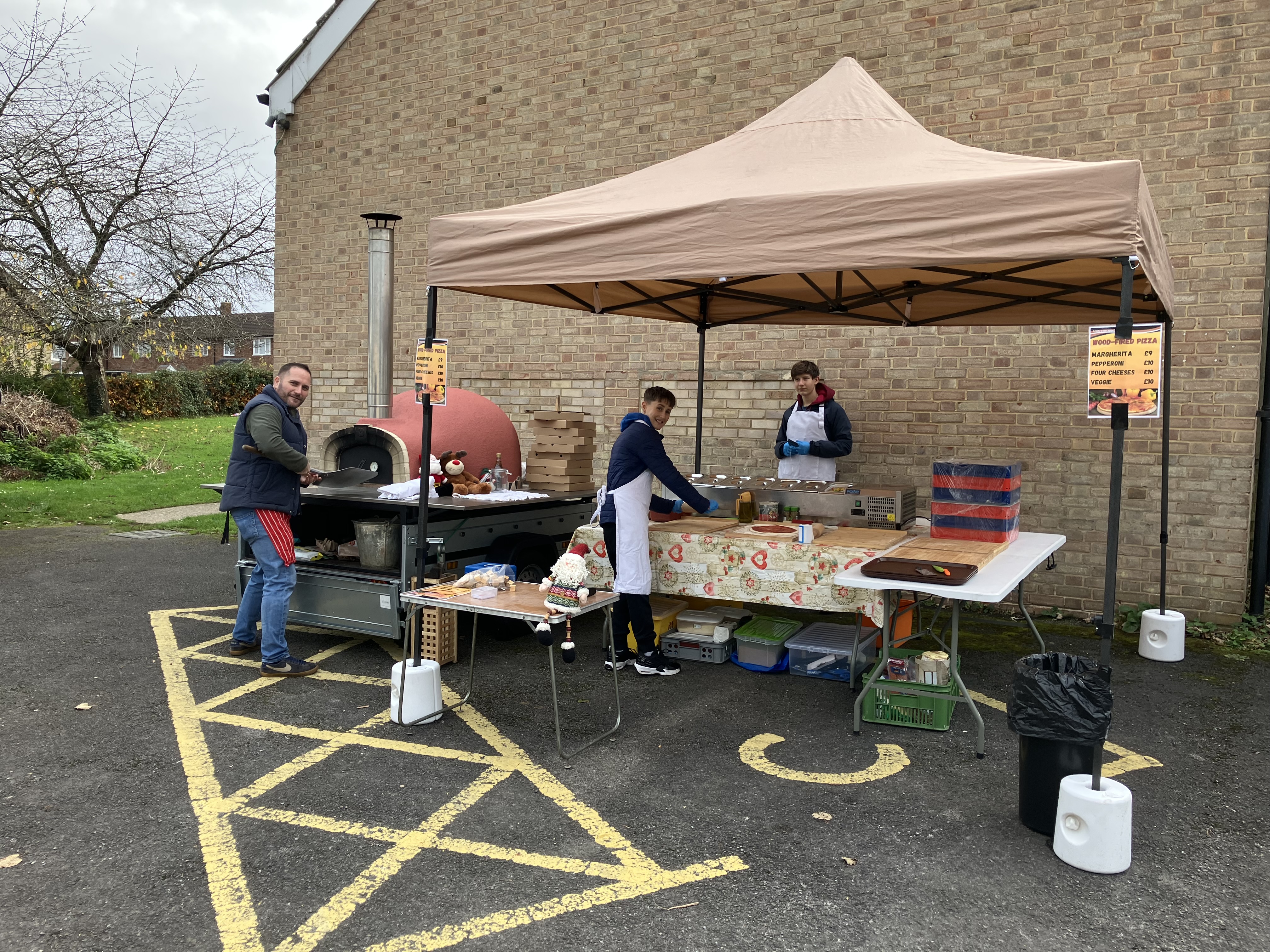 Outdoor mobile wood-fired pizza stand with staff preparing pizzas near brick building
