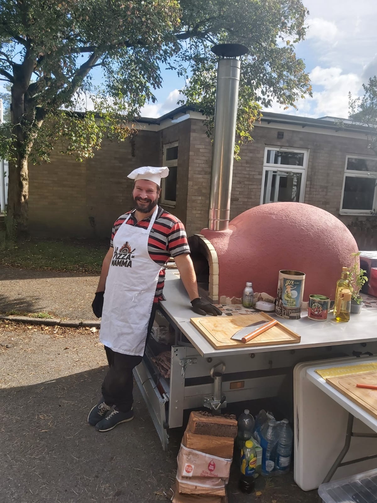 Pizza chef in hat and Pizza di Mamma apron beside red wood-fired oven outdoors