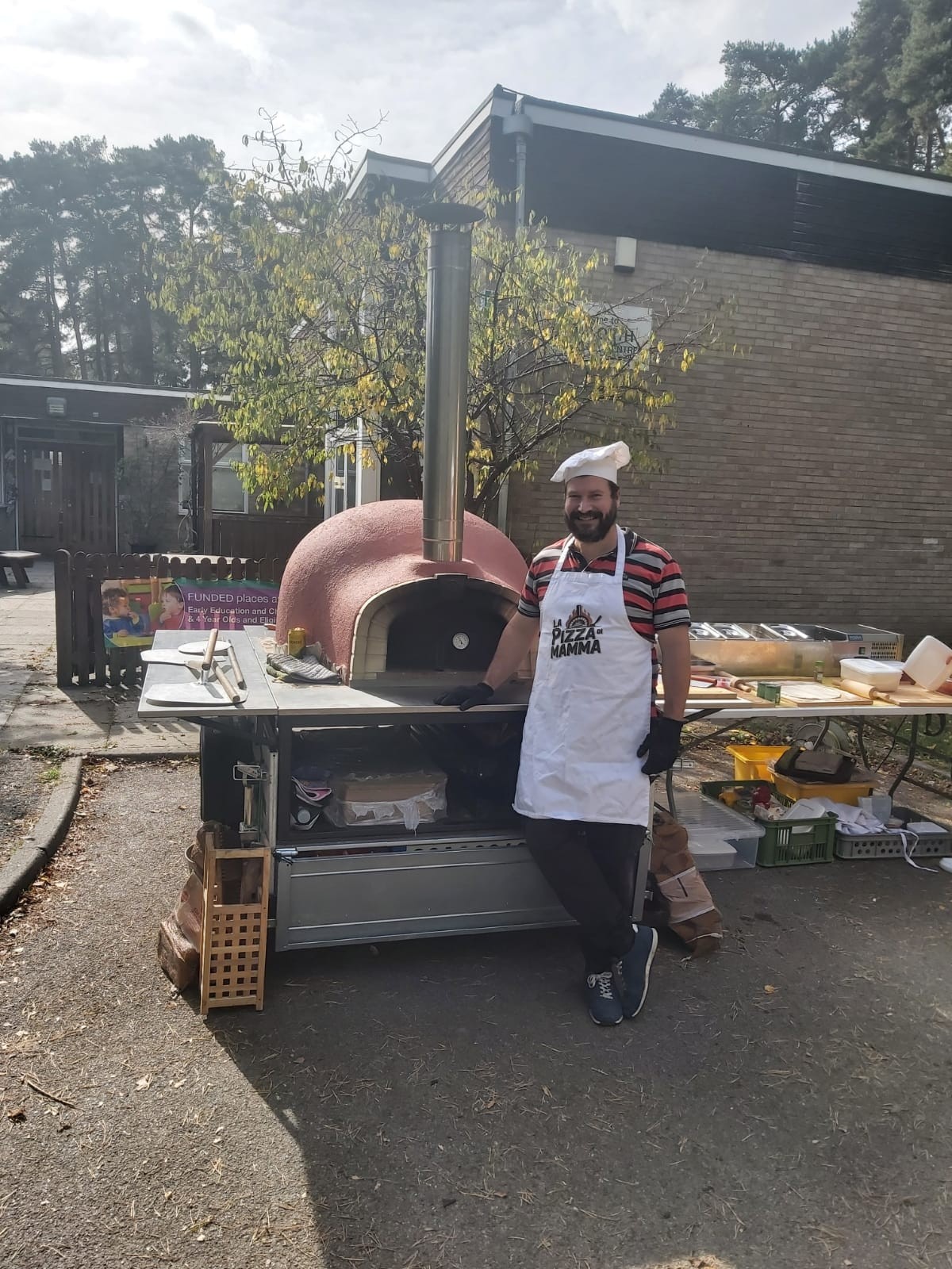 Pizza chef in Pizza di Mamma apron beside wood-fired pizza oven with outdoor backdrop